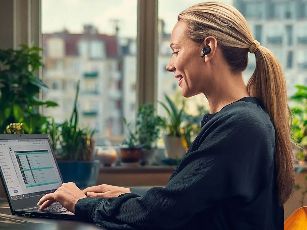 Een vrouw met blond haar werkt thuis achter de laptop en draagt de EPOS Adapt E1 Scandinavian Black draadloze Bluetooth oortjes voor professionals