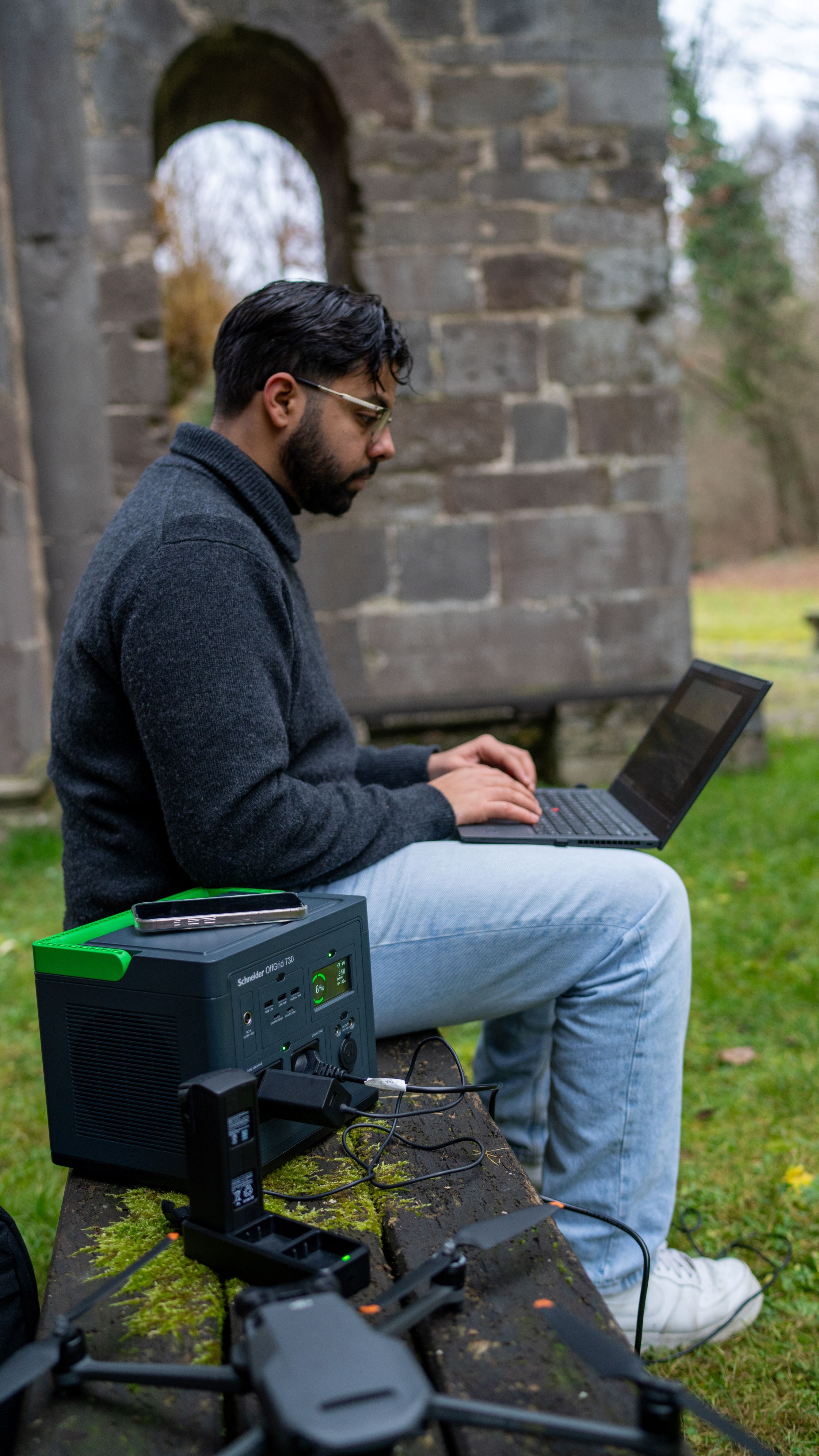 Een persoon zit buiten op een bankje met een laptop en een drone en het APC PPS730-GR draagbare oplaadstation voor stroom