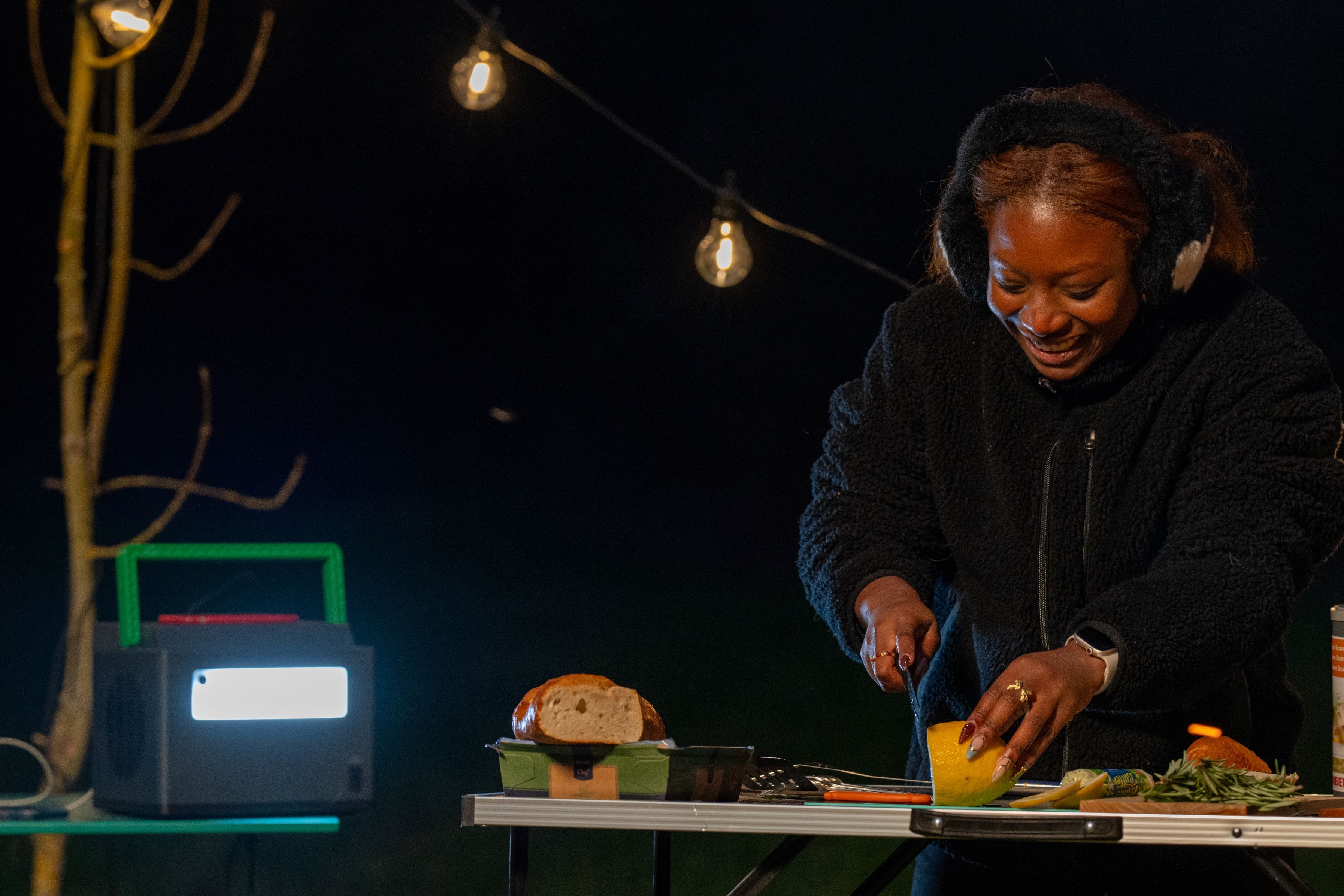 Het APC PPS730-GR draagbare oplaadstation staat op een campingtafel en daarnaast bereidt een persoon eten